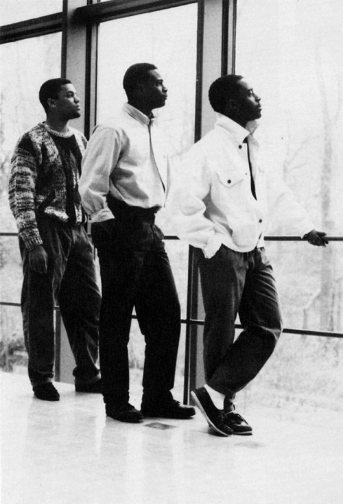 three male students look out a campus window while standing in a line